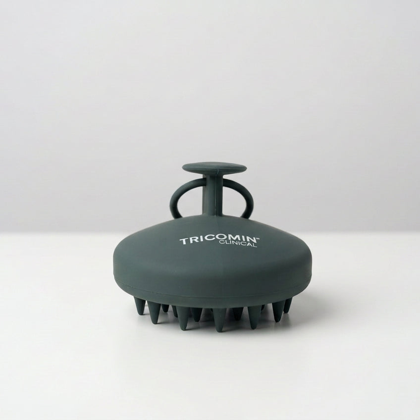 Tricomin - Scalp Massager - Front - A dark grey-green silicone scalp massager by Tricomin Clinical, featuring a circular head with cone-shaped bristles and a handle, on a clean white and light gray background.